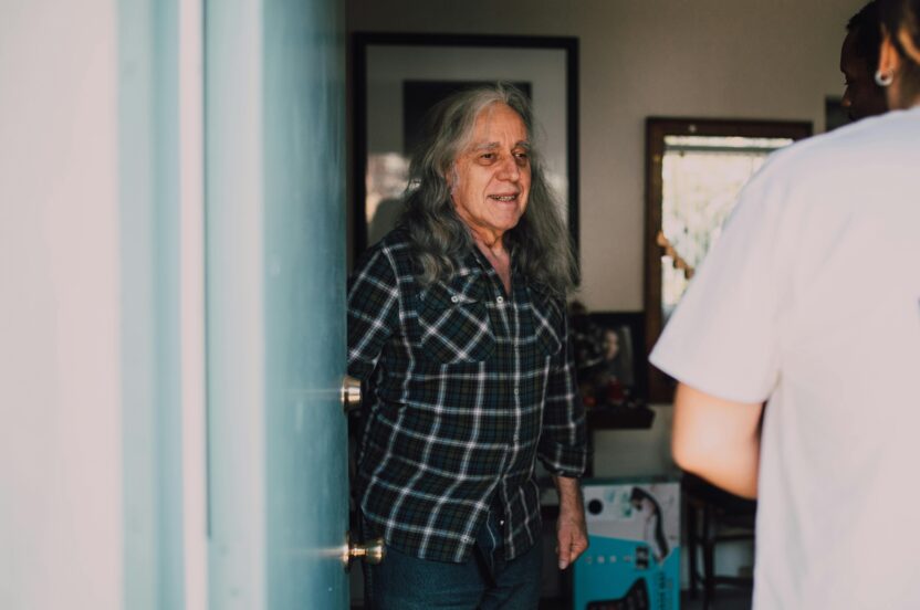 Older man with long grey hair opening his front door to a community health worker during a home visit.